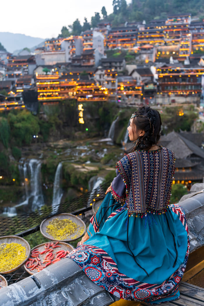 Turista vestida con ropa tradicional china admirando la vista panorámica del pueblo flotante de Furong y la cascada al anochecer.