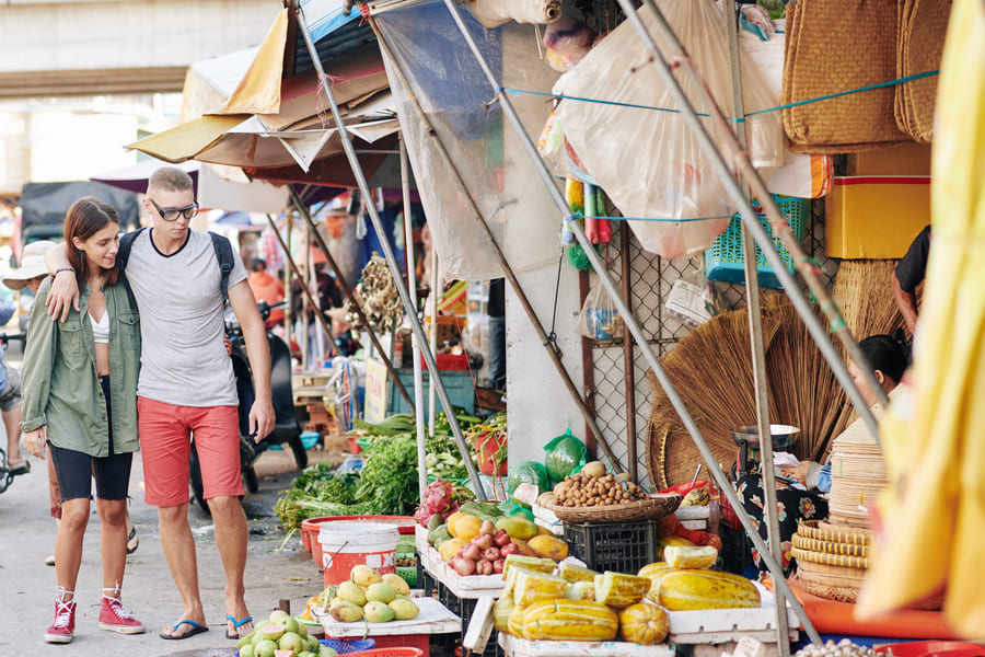 Jóvenes viajeros explorando un mercado tradicional en China, rodeados de puestos con frutas tropicales y productos locales.